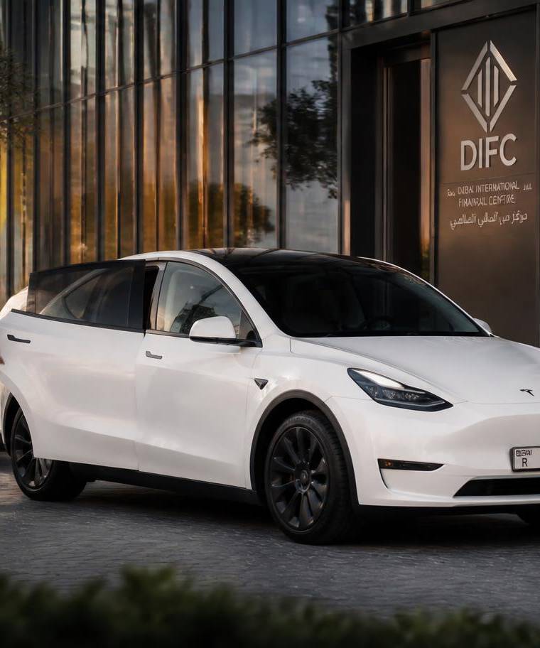 White Tesla Model Y chauffeur car with professional driver outside a DIFC office tower in Dubai at golden hour
